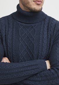 Pull en maille bleu marine à col montant, présentant des motifs complexes de torsades et de diamants, avec des poignets et un col côtelés pour une texture supplémentaire.