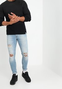 Suéter negro de canalé, jeans azul claro desgastados con rotos y zapatillas negras con acentos blancos, combinados con calcetines blancos.