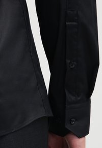 Maniche lunghe nere della camicia con polsini e bottoni al polso, cucitura laterale visibile e pantaloni neri su uno sfondo neutro.