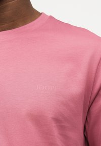 Κοντινό πλάνο ενός ατόμου που φορεί ροζ μπλούζα με στρογγυλή λαιμόκοψη και διακριτικό ανάγλυφο λογότυπο "JOOP! JEANS" στο στήθος.