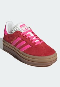 Czerwony zamszowy sneaker z różowymi akcentami i trzema białymi paskami. Posiada teksturowany biały język i gumową podeszwę w kolorze gumy. Model: Gazelle.