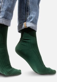 Zielone skarpetki o żebrowanym wzorze, wykonane z elastycznego materiału, z widocznymi kostkami i piętami, zestawione z niebieskimi dżinsami z podwiniętymi nogawkami.