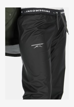 Black waterproof pants made of shiny, smooth fabric with an elastic waistband. Features the logo "SWEDEMOUNT" and mountain graphic in white.