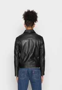 Persona con capelli ricci che indossa una giacca di pelle nera e jeans blu, in piedi di fronte a uno sfondo bianco semplice.