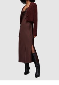 Hnědé saténové šaty s bočními rozparky doplněné vínově červeným krátkým kardiganem. Černé vysoké kozačky pod kolena dokončují outfit.
