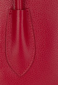 Detail einer roten Lederhandtasche, die die strukturierte Oberfläche, glatte Akzente und präzise Nähte hervorhebt und die einzigartige Form sowie Designelemente betont.