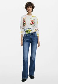White long-sleeve sweater features colorful floral patches; paired with blue flared jeans. The outfit is completed with black footwear.