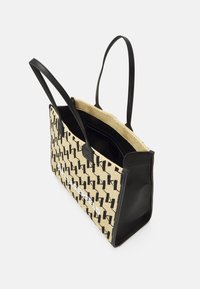 Stor tote-väska med svarta läderhandtag, beige vävd kropp med svarta geometriska mönster och en vit tryckt logotyp på framsidan.