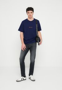T-shirt in cotone blu navy con testo ricamato arancione, abbinata a jeans in denim grigio e sneakers bianche con strisce nere e dettagli grigi.