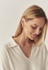 Cercei argintii cu design geometric, având un finisaj polişat. Modelul poartă un pulover crema cu decolteu în V și are părul drept, blond.