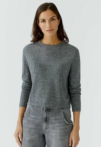 Frau mit dunklen Haaren, die einen grauen Strickpullover und hellgraue Jeans trägt und vor einem schlichten weißen Hintergrund steht.