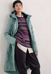 Lange turquoise pufferjas met een capuchon, gelaagd over een meerkleurige gestreepte trui en een wit shirt, gecombineerd met donkere broek. Glad textuur.
