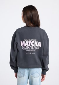 Donkergrijze sweatshirt met oversized pasvorm, met een roze tekstgrafiek op de achterkant: "New York City MATCHA MORNINGS" en aanvullende tekst eronder.