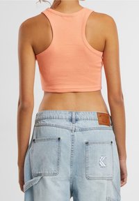 Žena nosící broskvový zkrácený tílko s racerback a světle modré džíny s vysokým pasem, které mají zadní kapsy a koženou značkovou nášivku.