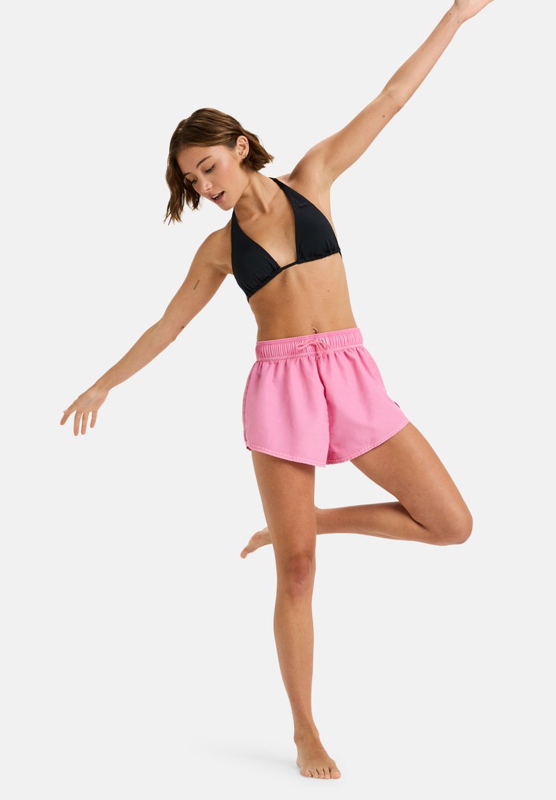 Schwarzes Triangel-Bikini-Oberteil kombiniert mit rosa Shorts mit Kordelzug, die einen lockeren Schnitt und ein leichtes Material aufweisen. Das Model posiert auf einem Bein.