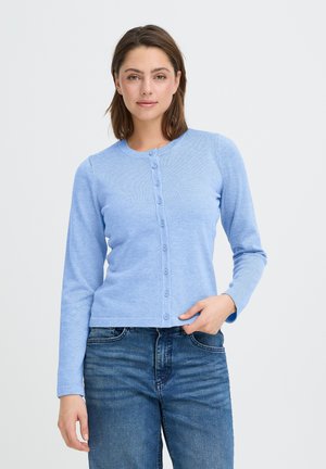 Kvinde iført en lyseblå knap-op cardigan og blå jeans, poserer med den ene hånd i lommen foran en ensfarvet hvid baggrund.