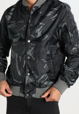 Mann trägt eine schwarz-grau camouflagefarbene Bomberjacke mit Druckknöpfen sowie gerippten Bündchen und Saum, Hände richten den Jackenbund.