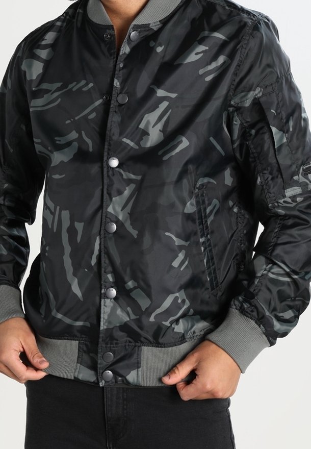 Homme portant une veste bomber camouflage noire et grise avec des boutons-pression et des poignets et ourlet côtelés, mains ajustant le bas de la veste.