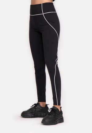 Mustat leggingsit korkealla vyötäröllä, joissa on valkoiset korostusviivat sivuilla. Kangas näyttää sileältä ja venyvältä. Yhdistetty paksuihin mustiin lenkkareihin.