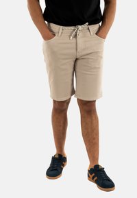 Shorts di cotone beige con vita regolabile, tasche anteriori e una superficie liscia, abbinati a scarpe da ginnastica blu navy con dettagli arancioni.