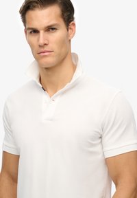 Weißes Poloshirt aus strukturiertem Stoff, mit einer Knopfleiste mit zwei Knöpfen und einem leicht erhöhten Kragen. Kurze Ärmel mit sauberen Kanten.