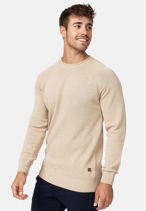 Beige knit sweater with long sleeves, round neckline, ribbed cuffs, and hem. Features a small logo patch near the bottom left.