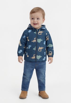 Niño pequeño sonriendo, vestido con una chaqueta azul con estampado de dinosaurios, vaqueros azules y botas color beige, de pie sobre un fondo blanco liso.