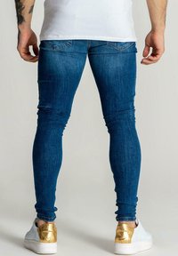 Vaqueros de mezclilla azul oscuro ajustados con un acabado texturizado, que cuentan con bolsillos traseros estándar. Combinados con zapatillas blancas y detalles en dorado.