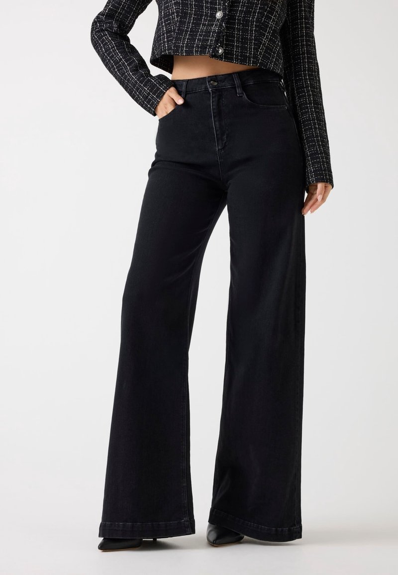 Femme portant un jean noir taille haute à jambes larges, des chaussures noires pointues et une veste courte à carreaux noir et blanc.