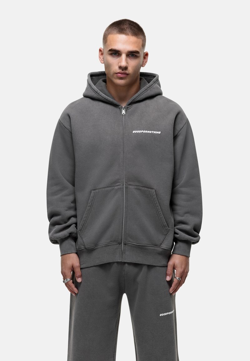 Sweatshirt à capuche gris zippé en matériau doux, avec une poche avant et un logo imprimé sur la poitrine gauche. Un pantalon de survêtement assorti est visible.