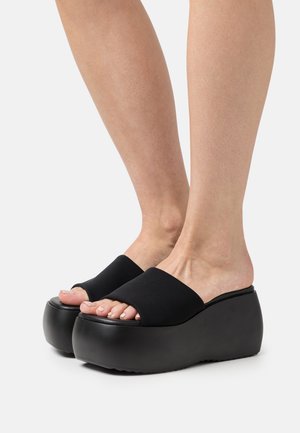 Gambe che indossano sandali con zeppa neri a pianta larga e punte aperte, su uno sfondo bianco uniforme.