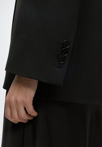 Gros plan d'une main reposant près de l'ourlet d'une manche de blazer noir avec trois boutons noirs sur le poignet.