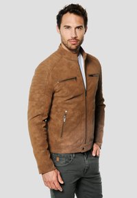Veste en cuir marron à coupe ajustée, avec des détails matelassés sur les épaules, des poches zippées et un col haut. Texture lisse et finition brillante.