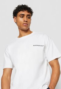 Hvit bomull T-skjorte med korte ermer, med et lite svart logo "MJGONZALES™" på venstre brystområde. Glatt tekstur og uformelt design.