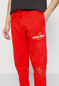 Røde sweatpants lavet af en bomuldsblanding; har grafiske tryk inklusive terninger og basketballmotiver, broderet tekst og en snor i taljen.