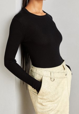 Mujer con un top negro ajustado de manga larga de canalé metido dentro de unos pantalones beige de cintura alta con detalle en anilla y manos en los bolsillos.