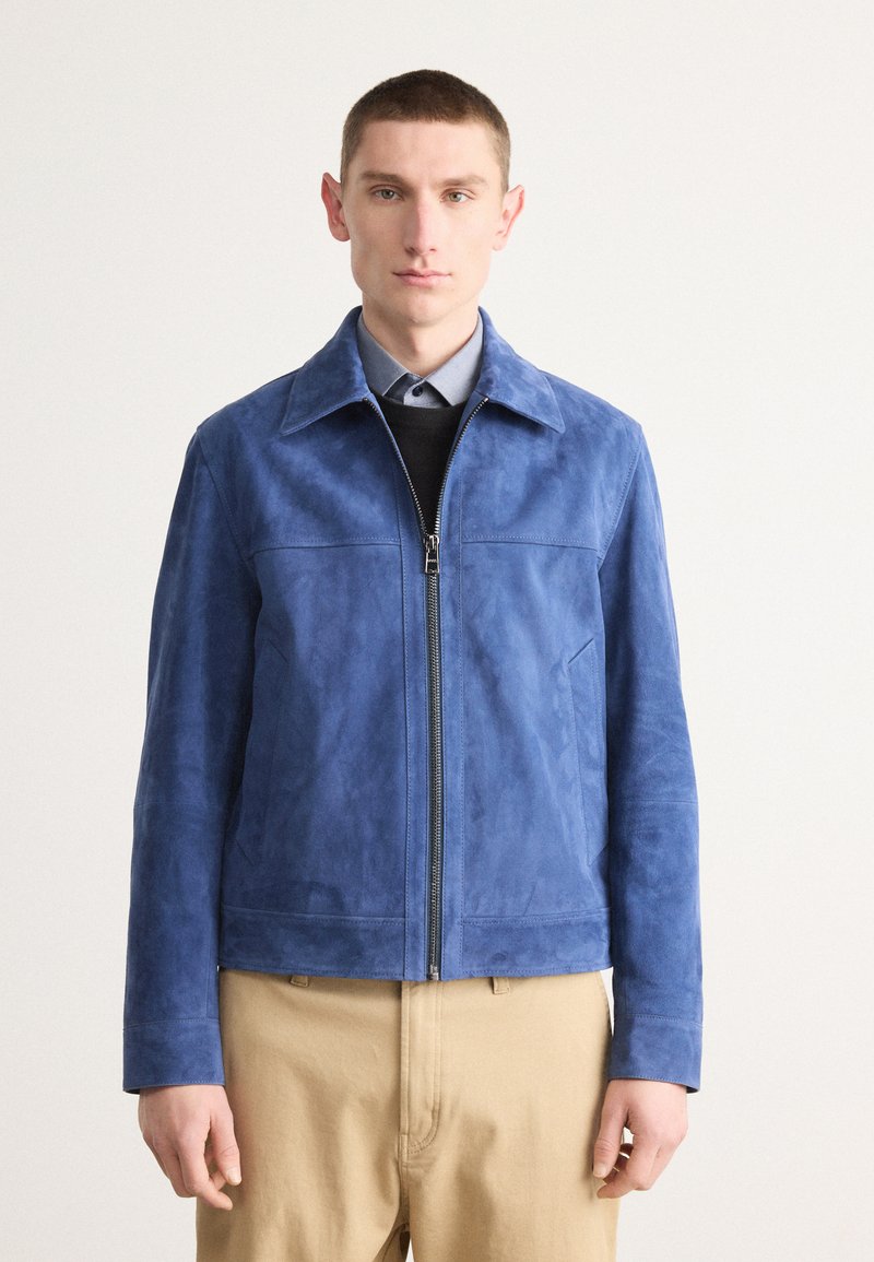 Μπλε σουέτ μπουφάν με φερμουάρ, μυτερό γιακά και δύο μπροστινές τσέπες. Λείος υφάσματος και προσαρμοσμένη εφαρμογή. Φορεμένο πάνω από ένα μαύρο πουλόβερ.