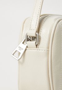 Crème neplederen tas met een rechthoekige vorm. Voorzien van een zilveren rits met een merkkel geschoven handvat en contrasterende witte stiksel details.
