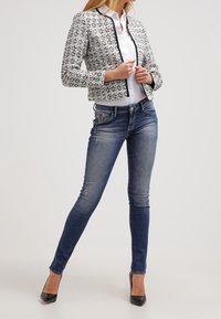 Chaqueta cortada de patrón en blanco y negro con un ribete negro sólido, usada sobre una camisa blanca, combinada con jeans azules y tacones negros.