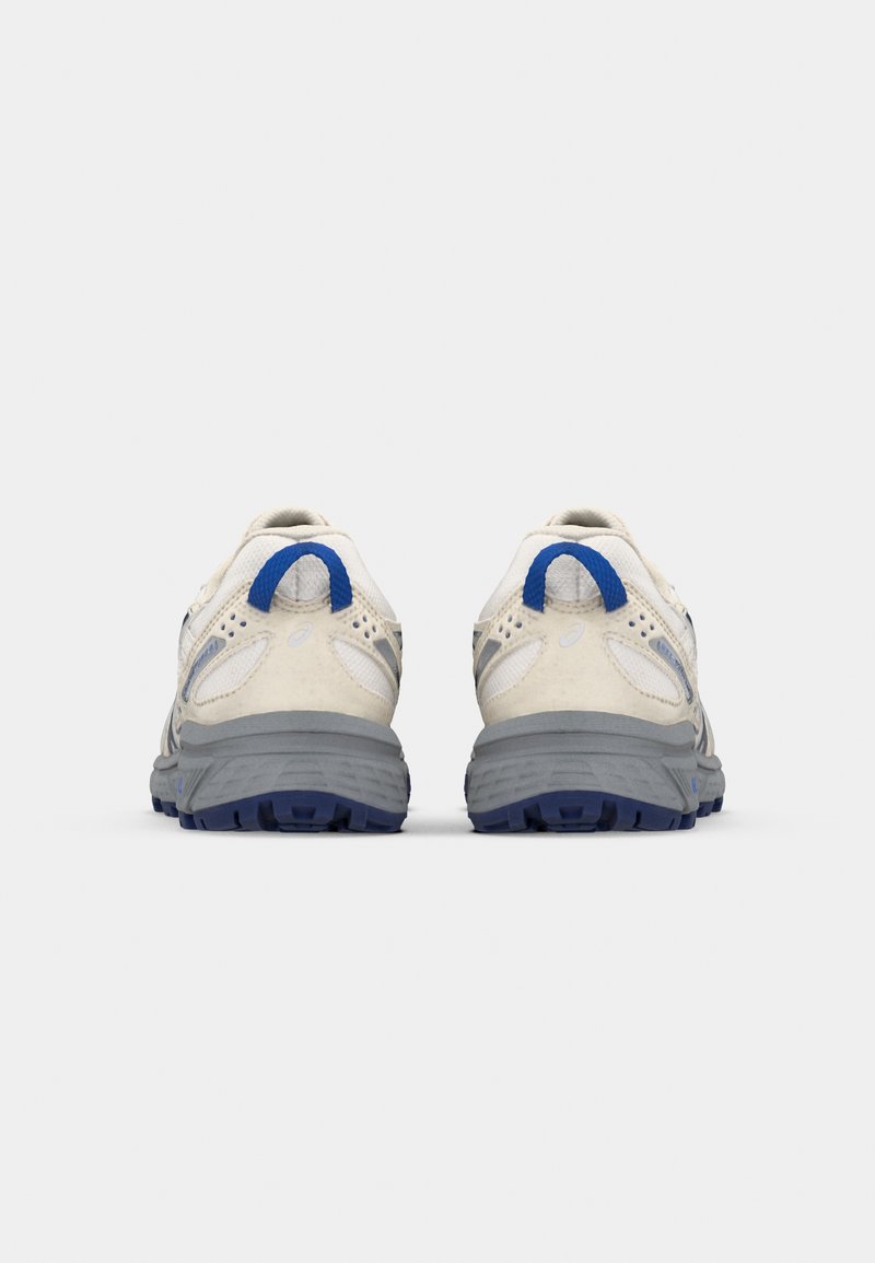 Zapatillas deportivas con parte superior de malla beige claro, acentos grises y una lengüeta azul. Media suela texturada en gris y suela exterior de goma negra.