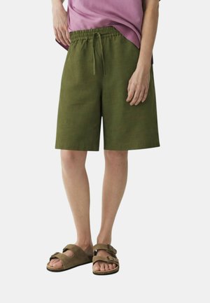 Persona che indossa pantaloncini verde oliva fino al ginocchio con vita con coulisse, camicia malva e sandali marroni con doppio cinturino.