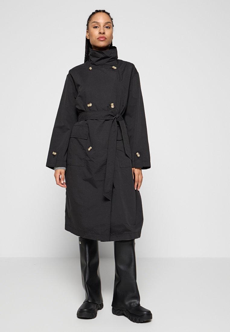 Schwarzer Trenchcoat mit hohem Kragen, goldenen Knöpfen und Taillengürtel. Verfügt über zwei Vordertaschen und einen geraden Schnitt. Getragen über schwarzen Stiefeln.