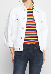 Chaqueta de mezclilla blanca con botones plateados, que presenta dos bolsillos en el pecho y mangas enrolladas, llevada sobre una blusa de rayas multicolores y jeans oscuros.