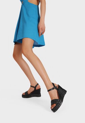 Donna che indossa un vestito blu e sandali con zeppa neri con cinturini alla caviglia, in piedi con un piede leggermente sollevato.