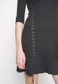 Gros plan d'une robe en maille côtelée gris foncé à manches 3/4 avec un détail à lacets sur le côté se terminant par des liens à embouts métalliques.