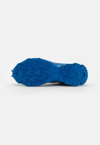 Semelle de chaussure de sport bleue avec un motif de grip texturé, présentant des crampons hexagonaux surélevés et une section centrale lisse. Accent rouge visible.