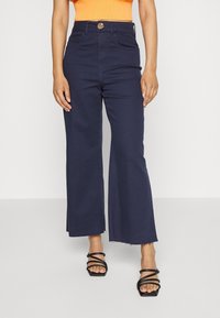 Bredbeniga mörkblå jeans med hög midja, framfickor, knappstängning och råa kanter vid fållen, i kombination med svarta sandaler med remmar.