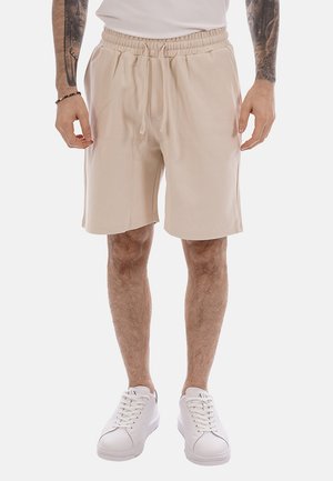 Uomo con pantaloncini beige a vita elastica e scarpe da ginnastica bianche, in piedi con le braccia rilassate lungo i fianchi, tatuaggi visibili sugli avambracci.