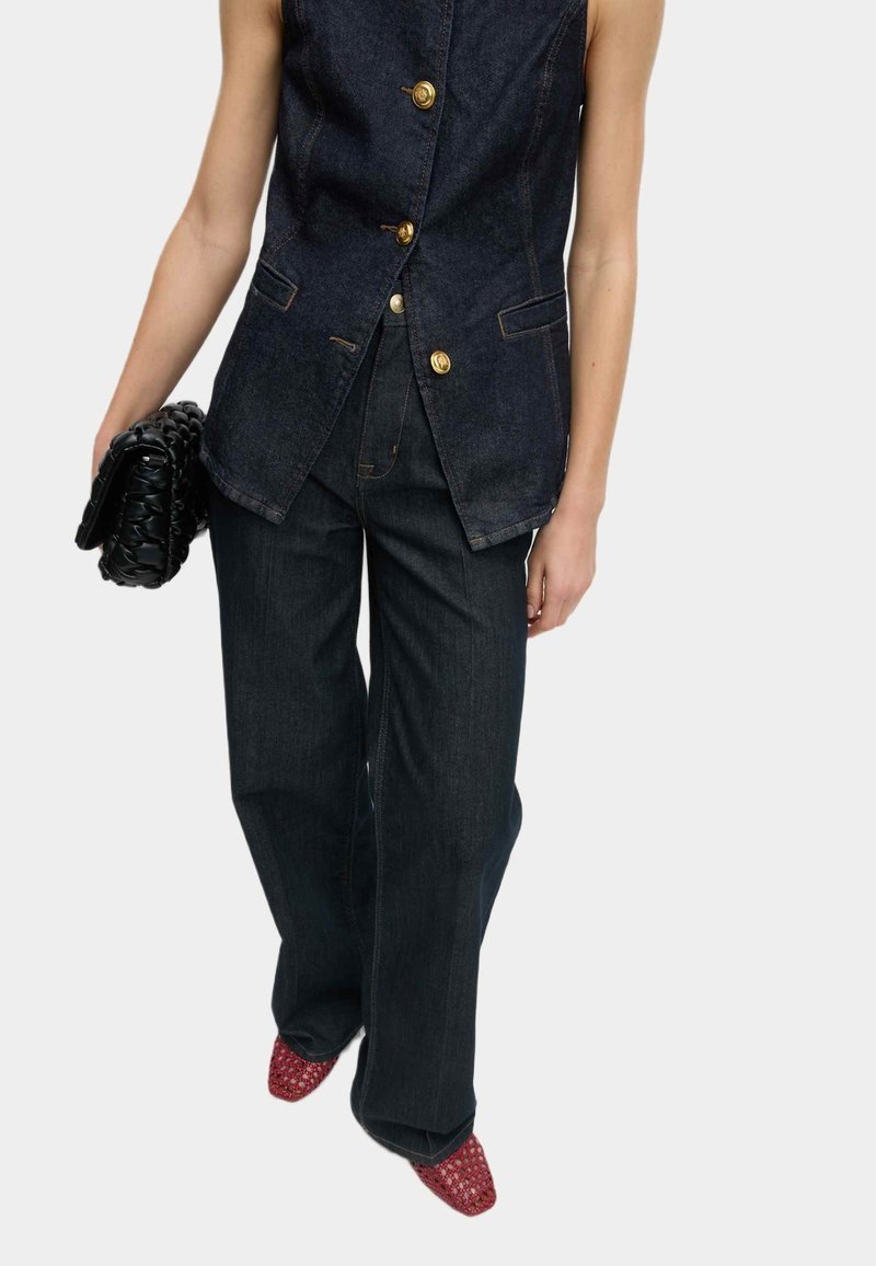 Femme portant un gilet sans manches en denim foncé avec des boutons dorés, un jean large assorti, tenant une pochette noire tressée et des chaussures rouges pointues.
