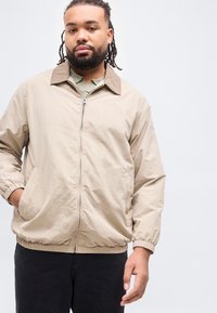 Άνδρας με γενειάδα και κράνη, ντυμένος με μπεζ μπουφάν με φερμουάρ πάνω από πράσινο πουκάμισο και μαύρο παντελόνι, στέκεται μπροστά σε απλό λευκό φόντο.